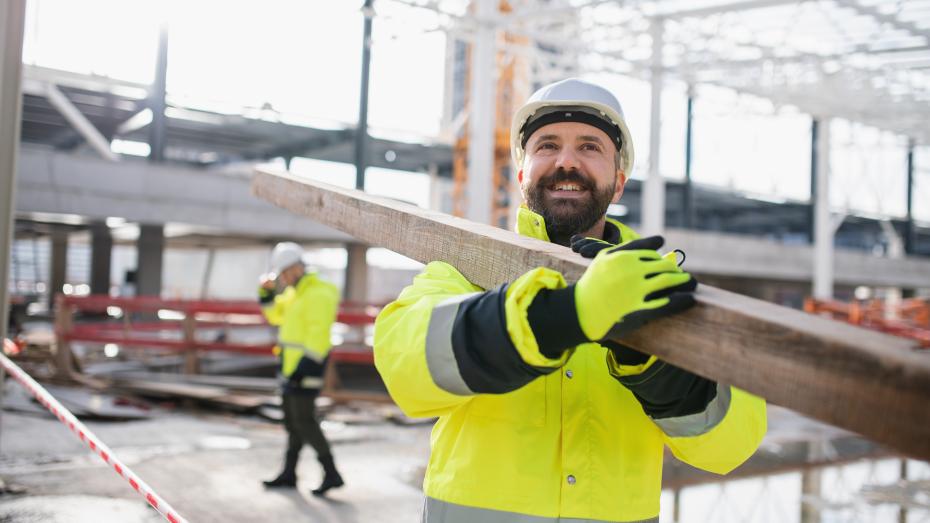 To industriarbeidere  i gule arbeidsjakker bærer treplanke på industriplass med stilas og stålkonstruksjoner. Foto Foto
