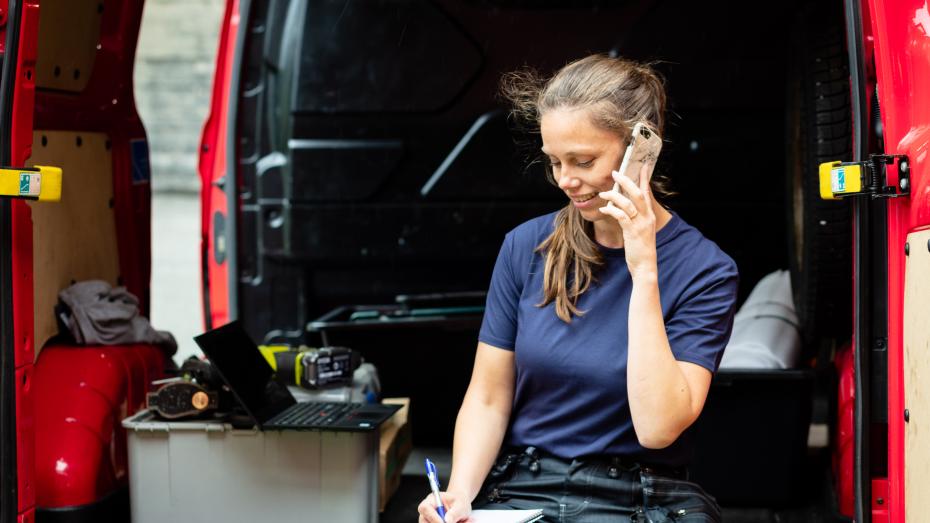 En kvinnelig håndverker sitter bak i en rød firmabil og snakker i telefonen. Foto