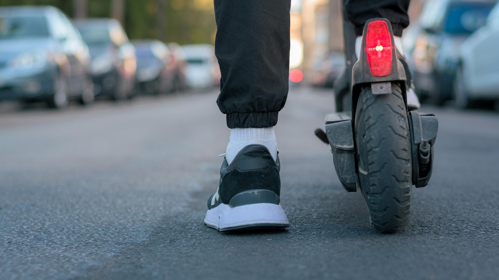 Nærbilde av person som triller en elsparkesykkel langs en bygate, sett bakfra.
