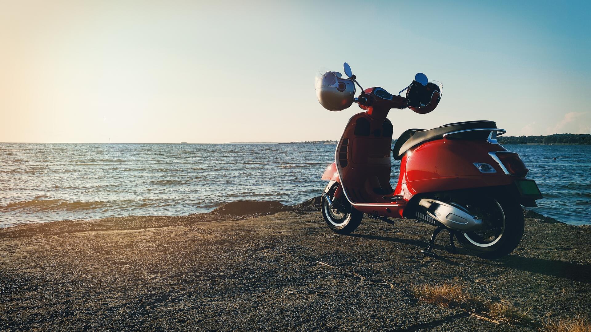 Rød scooter parkert ved sjøen i solnedgang.