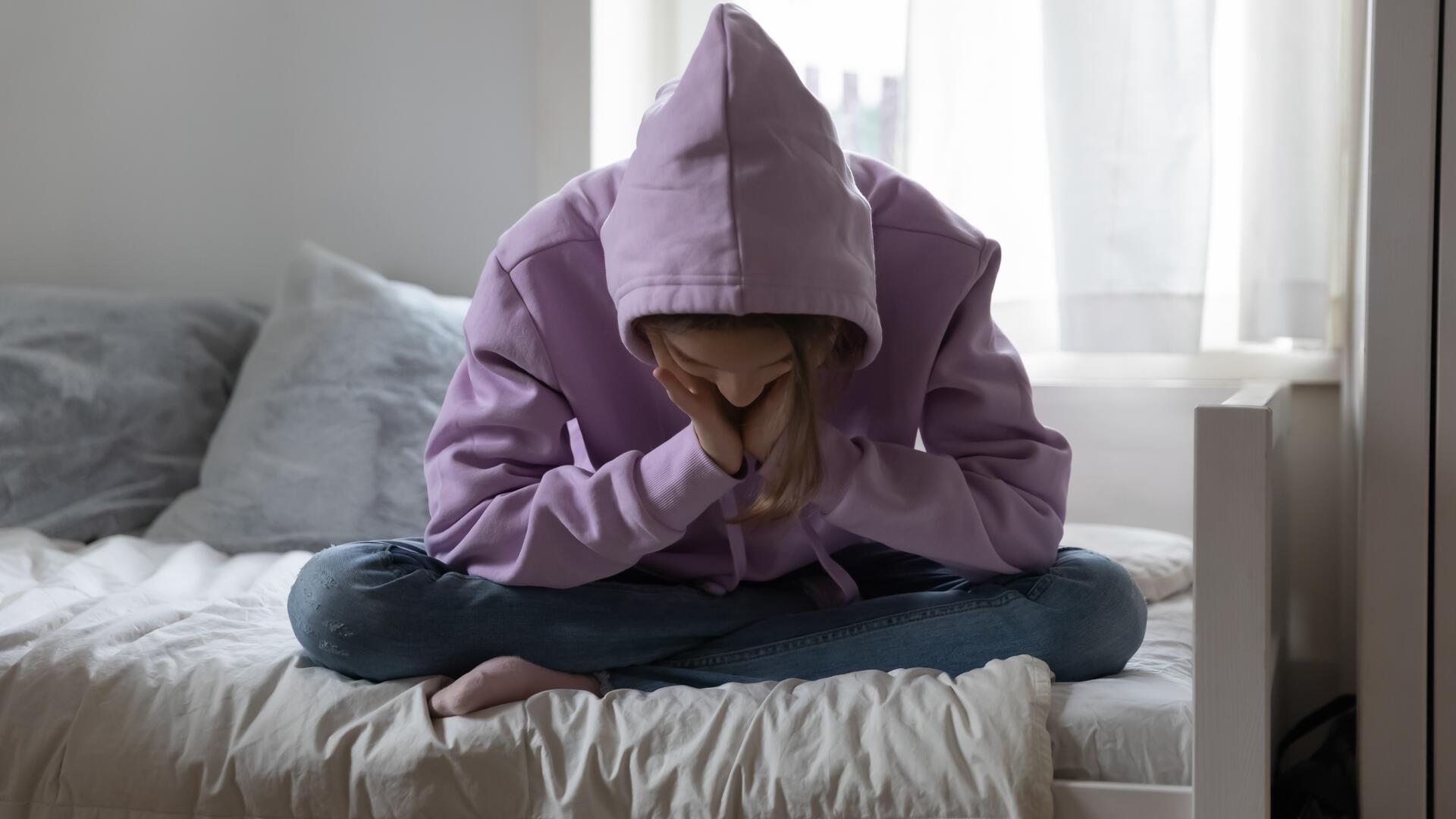 Ung person i hettegenser sitter nedstemt på en seng.