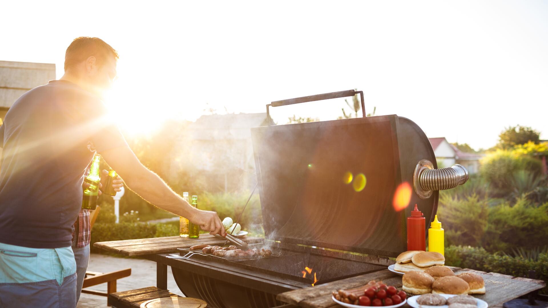  En mann griller kjøtt på en stor kullgrill i kveldssolen, omgitt av grilltilbehør som pølser, brød, tomater og dressinger.