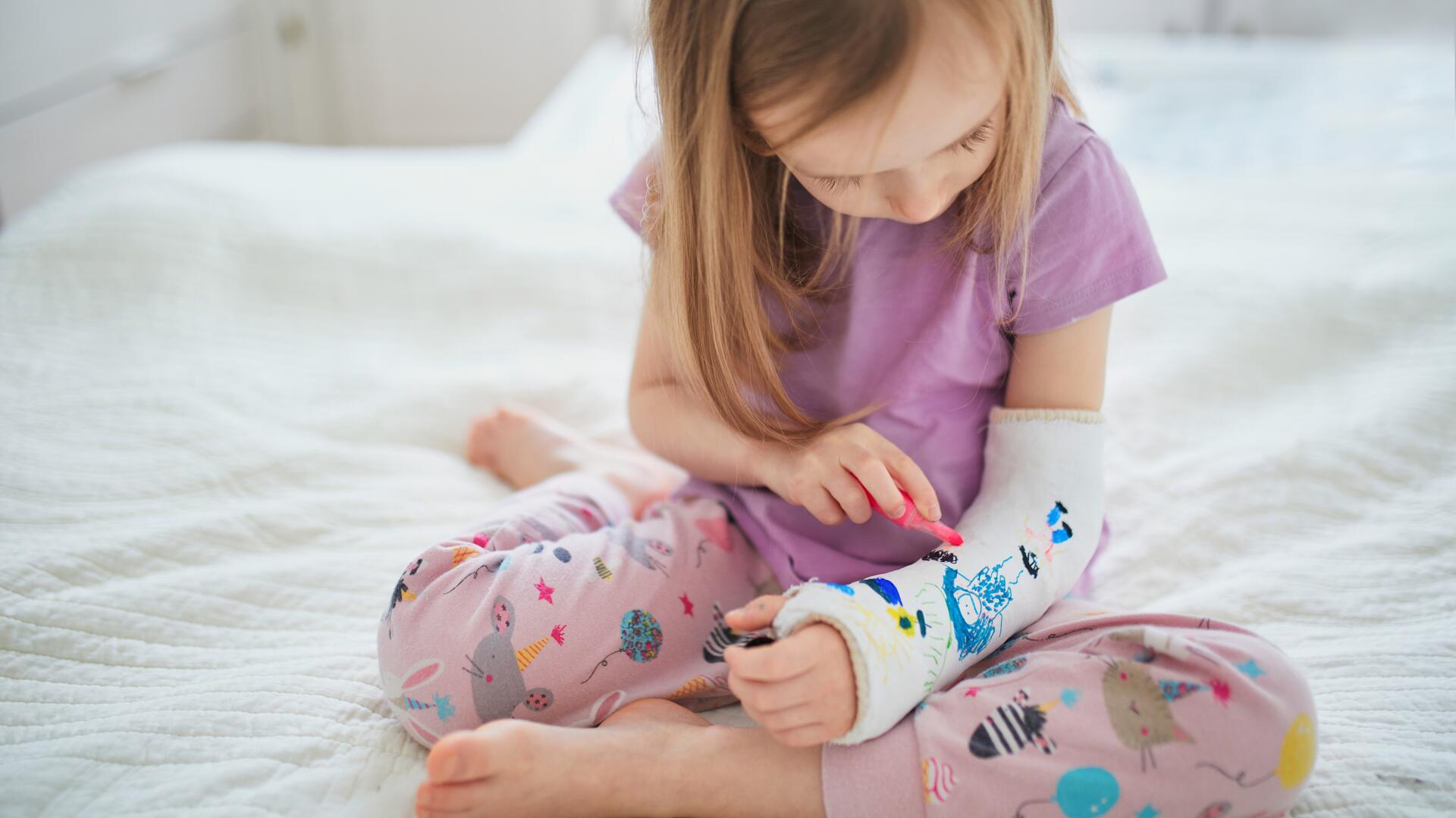 Liten jente med gipset arm sitter på sengen og tegner på gipsen sin.
