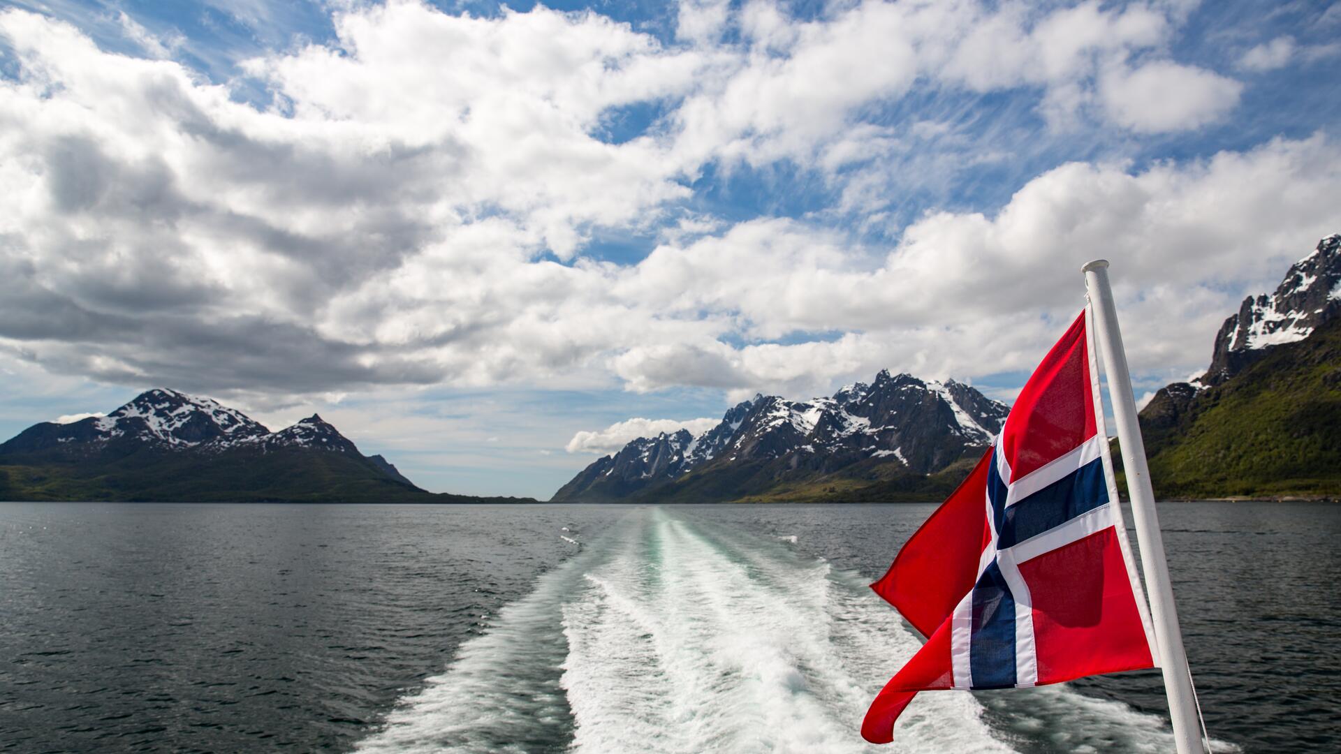 Norsk flagg vaier bak en båt i fart gjennom norsk fjordlandskap, med fjell og skyer i bakgrunnen.