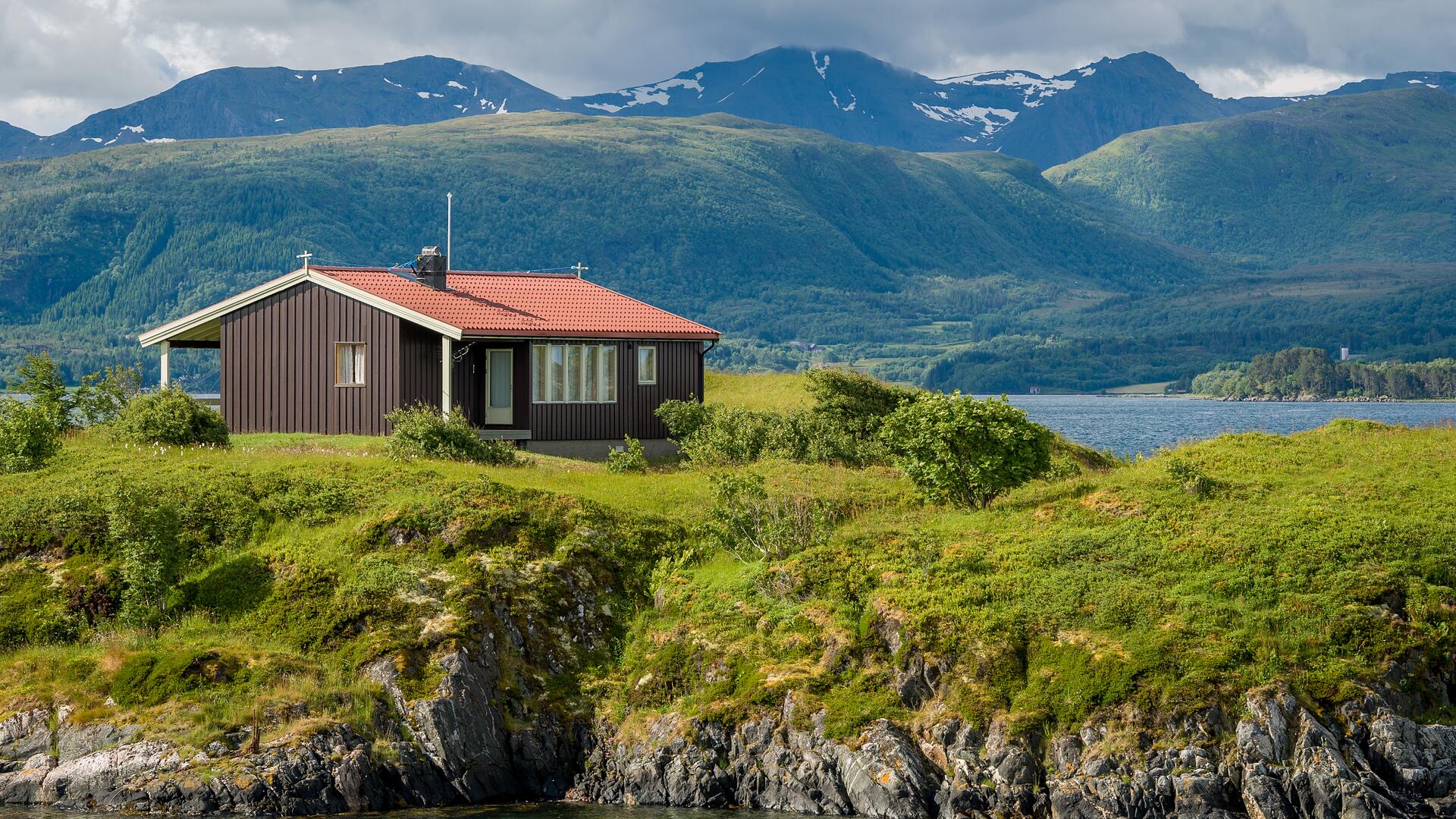 Hytte ved fjordlandskap omgitt av grønne åser og fjell, med vann i forgrunnen og skyet himmel.