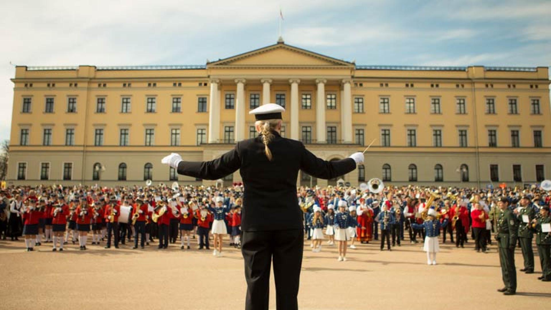 Dirigent leder et stort korps foran Slottet i Oslo på en solrik dag.