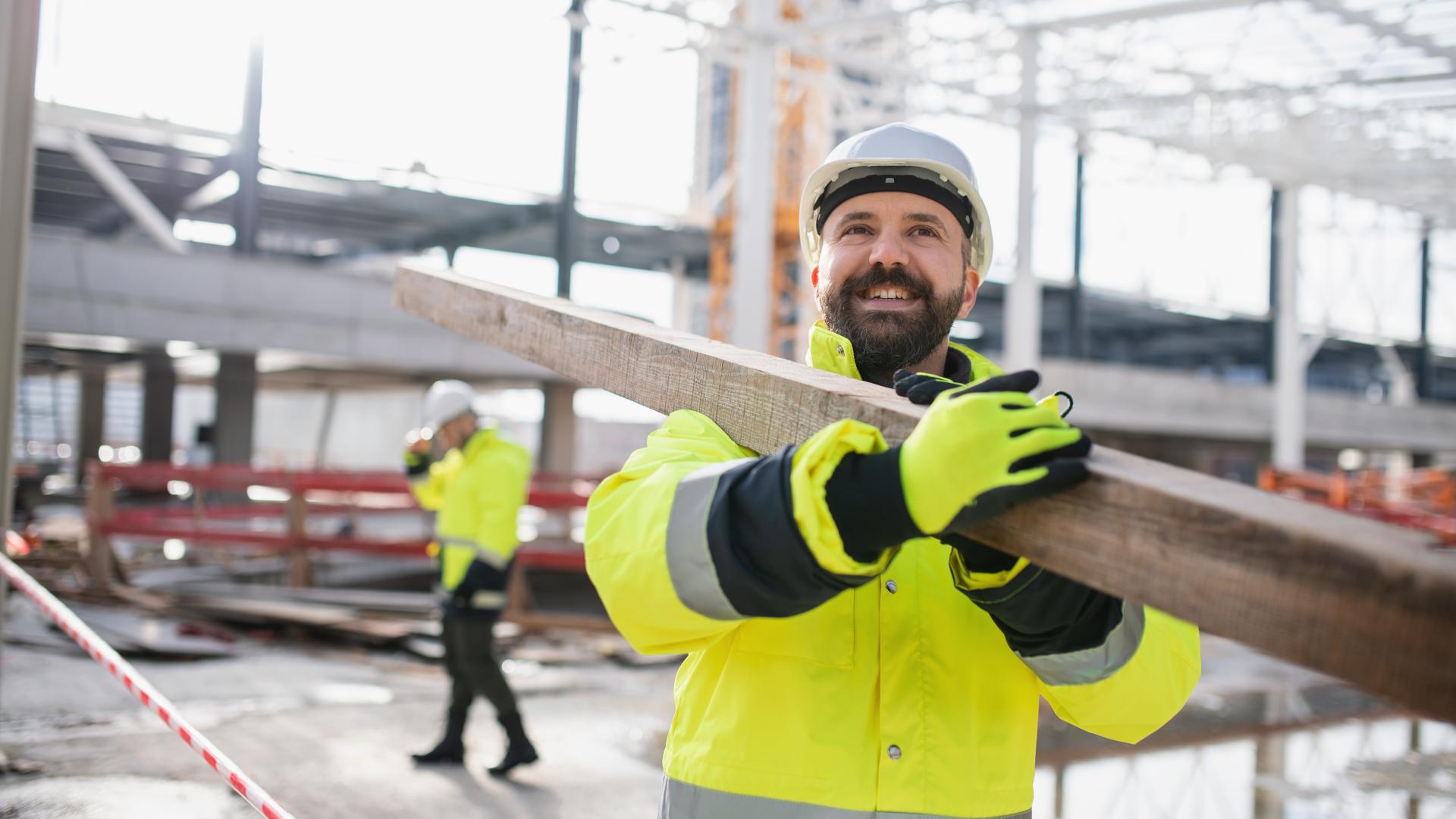 To industriarbeidere  i gule arbeidsjakker bærer treplanke på industriplass med stilas og stålkonstruksjoner. Foto Foto