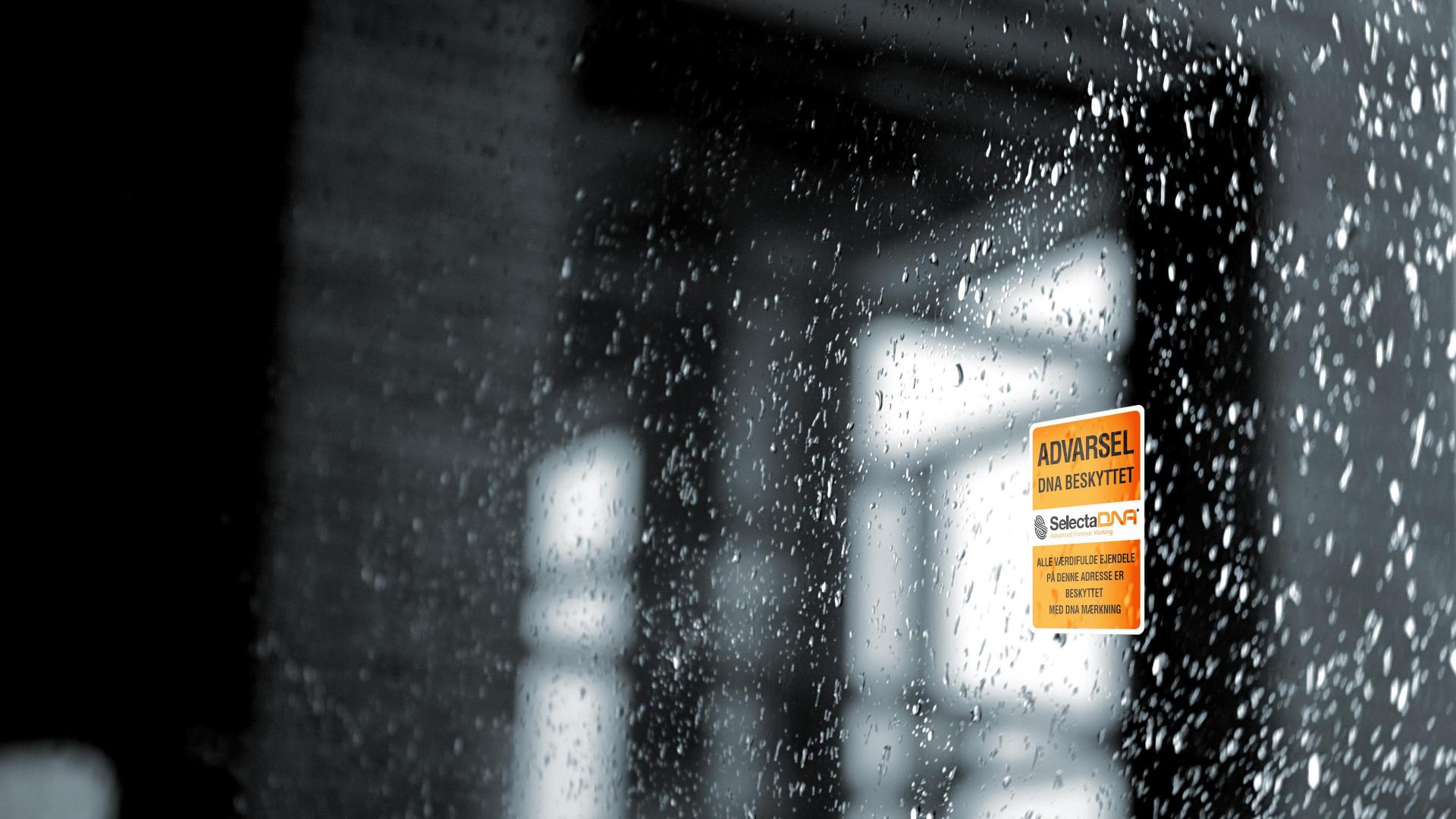Varselskilt fra SelectaDNA med teksten "ADVARSEL DNA-BESKYTTET", festet på en regnvåt vindusrute.