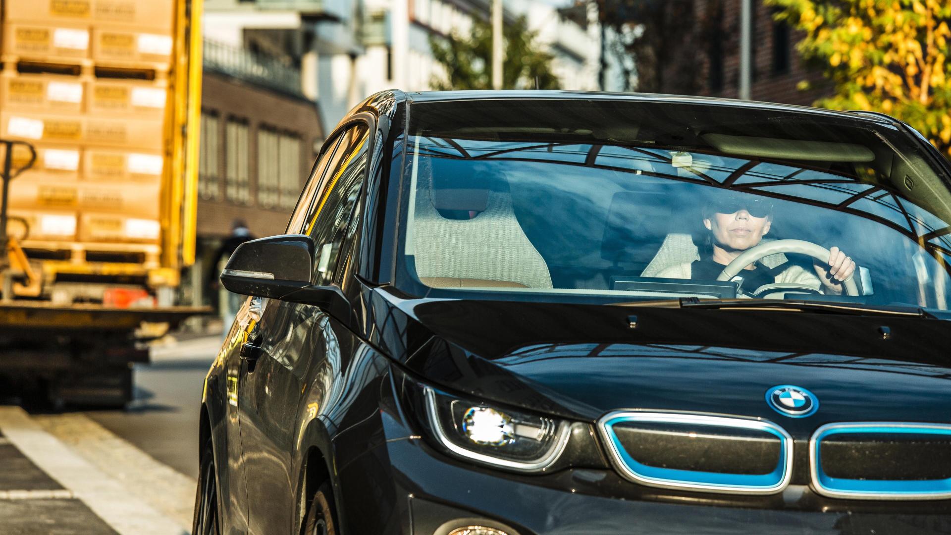 En fører kjører en svart BMW elbil i bytrafikk med lastebil i bakgrunnen.