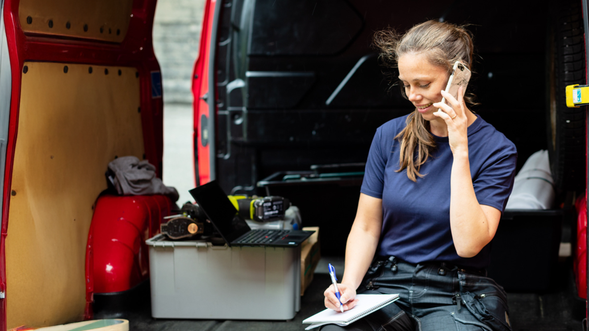 Kvinne sitter i arbeidsbilen og snakker i telefonen og noterer