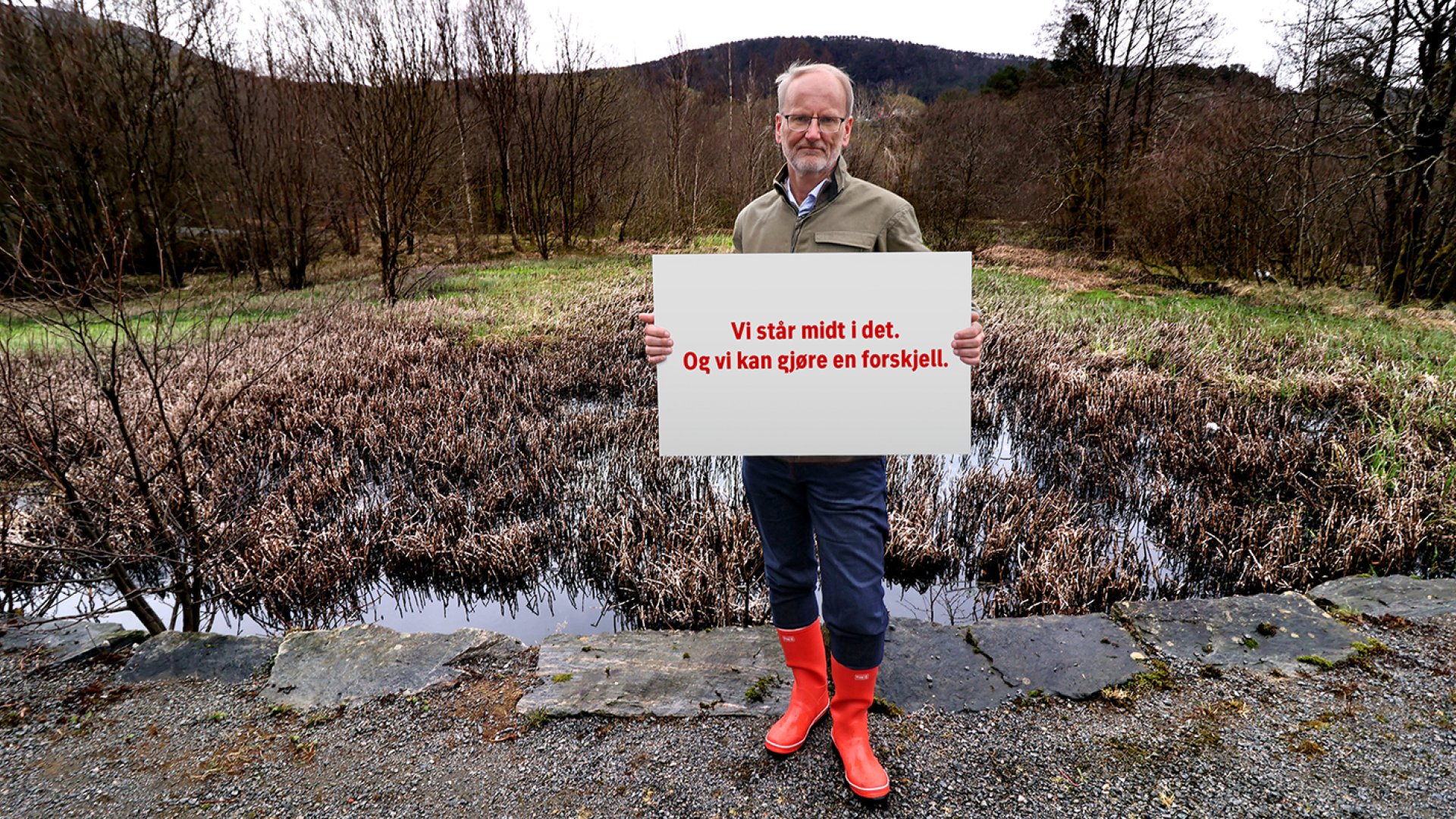 Person med røde gummistøvler står ved et vått naturområde og holder et skilt med teksten "Vi står midt i det. Og vi kan gjøre en forskjell."